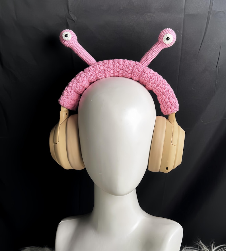 Op de afbeelding: Roze gehaakt hoofdband met twee cartoonogen, ontworpen om over koptelefoons te passen. Het hoofdband wordt gedragen door een wit mannequin.