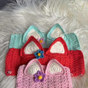 May include: Three crocheted headbands with white ears and a flower embellishment. The headbands are in shades of pink, red, and light blue.