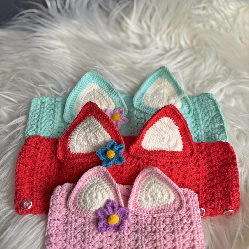 May include: Three crocheted headbands with white ears and a flower embellishment. The headbands are in shades of pink, red, and light blue.