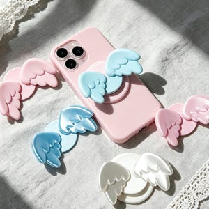 Può includere: Una custodia per smartphone rosa chiaro con un supporto per telefono a forma di ali d'angelo azzurre fissato sul retro. Attorno al telefono ci sono tre supporti circolari aggiuntivi con ali d'angelo nei colori rosa, azzurro e bianco.
