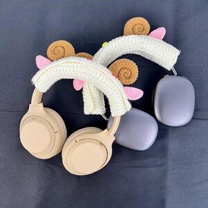 K&ouml;nnte beinhalten: Zwei Paar beigefarbene kabellose Kopfh&ouml;rer mit wei&szlig;en geh&auml;kelten Ohrensch&uuml;tzern mit braunen und rosa geh&auml;kelten Schafmotiven.