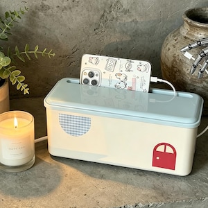May include: A rectangular cable management box with a light blue lid and a cream-colored base. The base features a blue and white checkered semi-circle and a small red door graphic. A smartphone with a white character-patterned case is inserted.