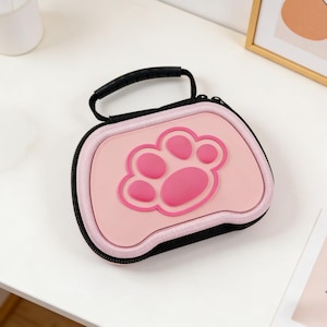 Könnte beinhalten: Eine rosa-schwarze Reißverschluss-Tragetasche mit schwarzem Griff. Die Tasche hat ein erhabenes, hellrosa Pfotenabdruck-Design auf der Vorderseite. Die Tasche ist wahrscheinlich für kleine Elektronik oder Zubehör.