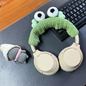 May include: White headphones with a green crocheted frog headband. The headband has two large white eyes with black pupils.