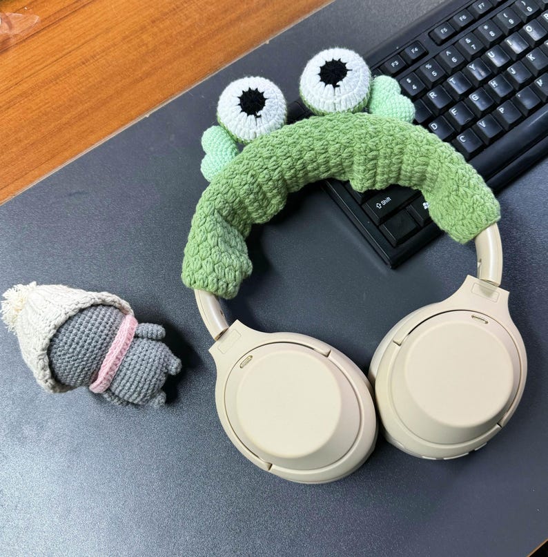 May include: White headphones with a green crocheted frog headband. The headband has two large white eyes with black pupils.