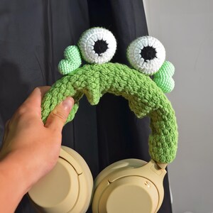 May include: Green crocheted headband with large white eyes and a green crocheted frog design. The headband is on a pair of beige headphones.