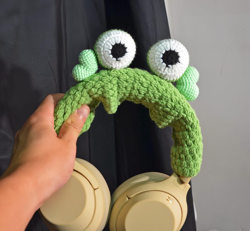May include: Green crocheted headband with large white eyes and a green crocheted frog design. The headband is on a pair of beige headphones.