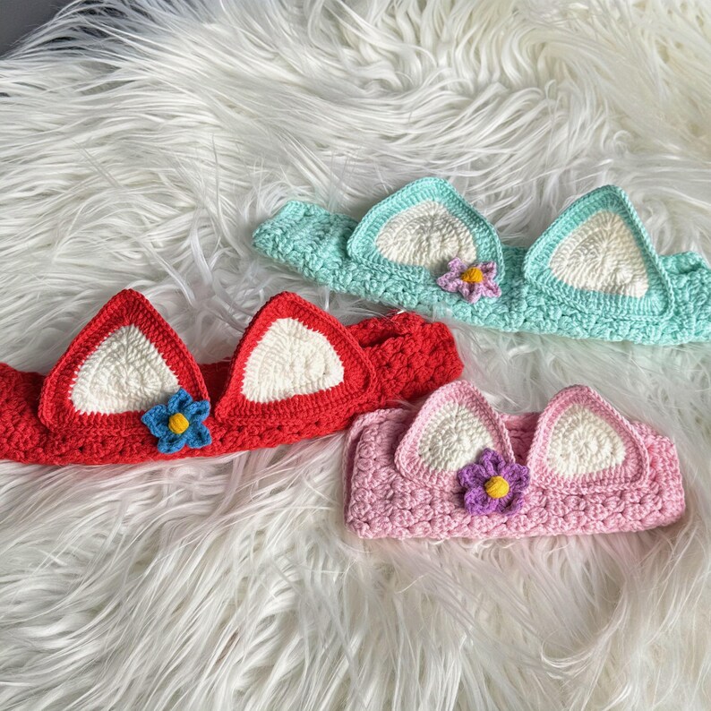 May include: Three crocheted headbands with cat ears and a flower detail. The headbands are in red, pink, and light blue.
