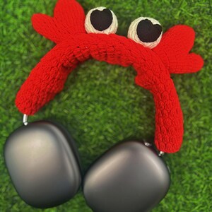 Op de afbeelding: Een rood gebreid hoofdband in de vorm van een krab met zwarte ogen en witte accenten. Het hoofdband wordt gedragen over een zwarte koptelefoon.