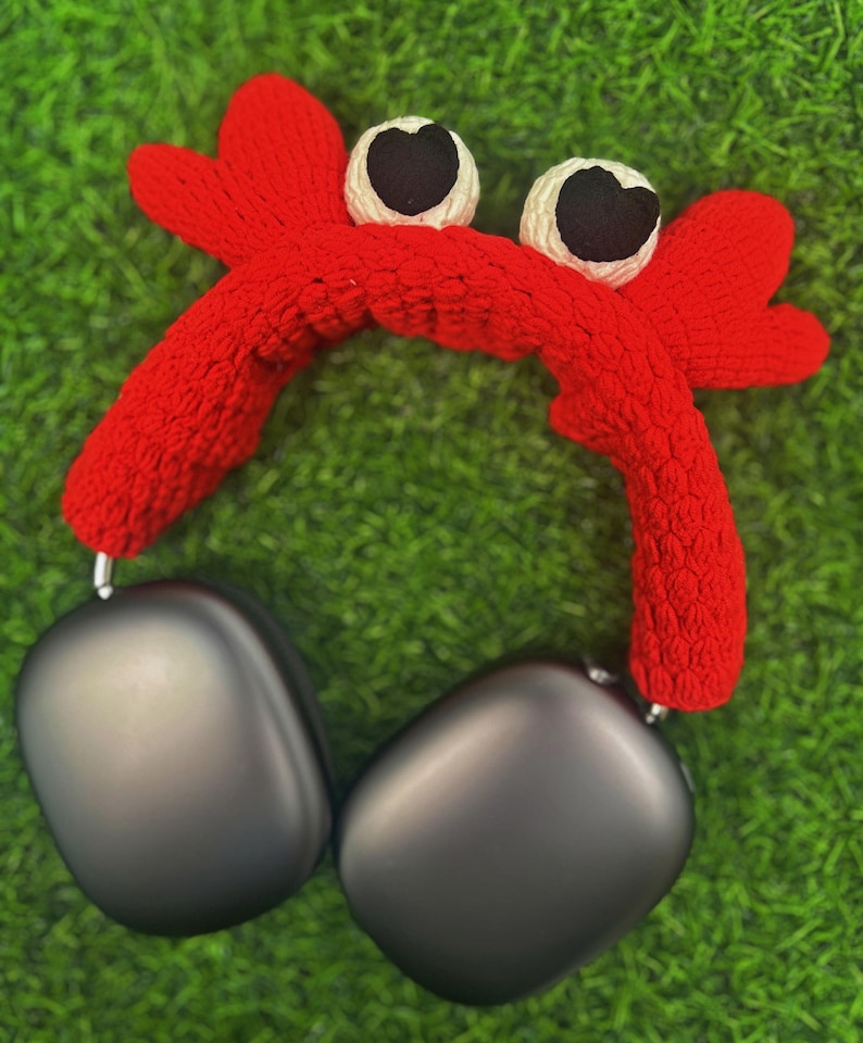 Op de afbeelding: Een rood gebreid hoofdband in de vorm van een krab met zwarte ogen en witte accenten. Het hoofdband wordt gedragen over een zwarte koptelefoon.