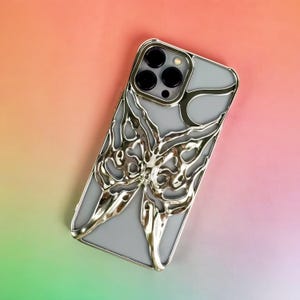 May include: A silver phone case with a butterfly design. The case has a metallic finish and a gray interior. The butterfly design is intricate and detailed, with a flowing, organic shape. The case is shown against a colorful gradient background.