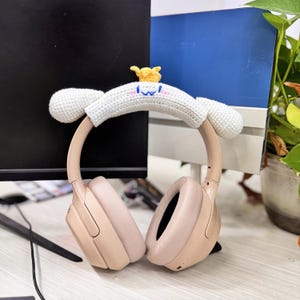 May include: Blush-colored over-ear headphones with a white crocheted headband cover. The headband cover has a cartoon design with a yellow crown and blue accents. The headphones are resting on a desk.