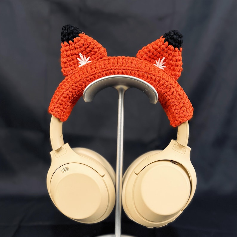 May include: Beige headphones with a crocheted fox ear headband. The headband is orange with black and white accents.