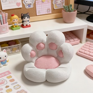 以下が含まれることがあります： 白とピンクの猫の足の形をした携帯電話スタンド。 スタンドは、ピンクのパッドとピンクの中央部を備えた、質感のあるふわふわした外観をしています。 スタンドは白い表面にあり、背景には他のデスクアクセサリーがあります。