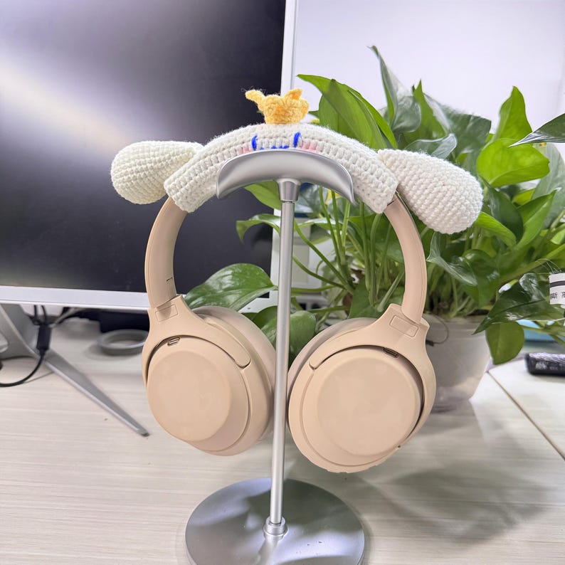 May include: A pair of beige wireless headphones resting on a silver stand. The headphones are adorned with a white crocheted cover shaped like a bunny, complete with ears and a small yellow crown. The stand has a circular base.
