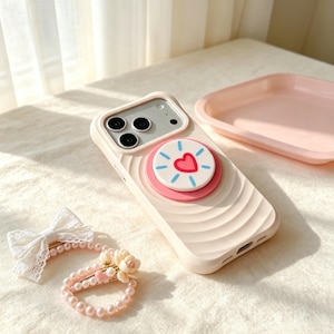 May include: A cream-colored smartphone case with a wavy texture and a circular phone grip featuring a red heart and blue rays. Beside it lies a hair accessory with a white lace bow, a string of light pink pearls, and small floral accents.