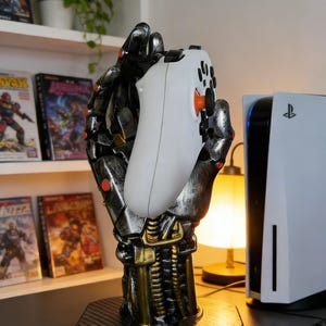 Peut inclure: Un support métallique en forme de main robotique tient une manette de jeu blanche avec des accents orange. Le support est argenté et doré, avec une base texturée. Une console PlayStation 5 est visible en arrière-plan.