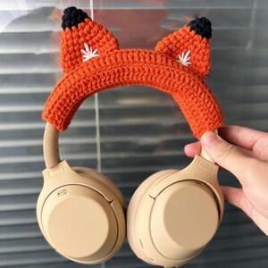 May include: Orange and black crocheted fox headphone cover for over-ear headphones. The cover has white embroidered details on the ears.