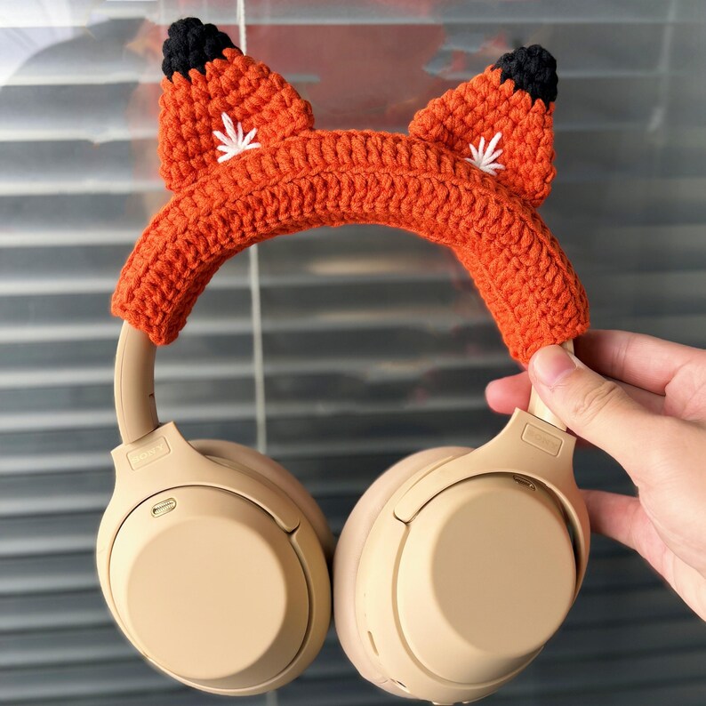 May include: Orange and black crocheted fox headphone cover for over-ear headphones. The cover has white embroidered details on the ears.
