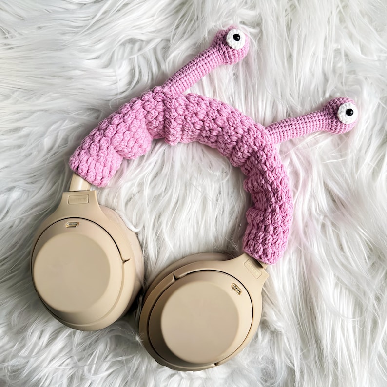 Op de afbeelding: Een beige koptelefoon met een roze gehaakt hoofdband. De hoofdband heeft twee kleine roze gehaakte ogen.