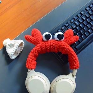 Op de afbeelding: Witte koptelefoon met een rood gebreid krabbenhoofdband. De hoofdband heeft twee grote zwarte ogen.