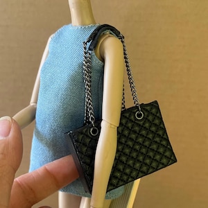 Puede incluir: Un pequeño bolso negro acolchado con una correa de cadena plateada. El bolso es de forma cuadrada y tiene un patrón de diamantes. El bolso lo lleva una persona que viste un vestido vaquero azul.