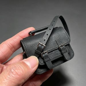 May include: A miniature black satchel bag featuring a textured surface, a top handle, and a long shoulder strap. The front flap is secured with two vertical straps that include silver-toned metal buckles and punched holes for adjustment.