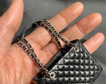 Könnte beinhalten: Eine schwarze Miniatur-Handtasche mit gesteppter Textur und einem silberfarbenen Kettenriemen, der mit schwarzem Material durchflochten ist. Die Tasche hat eine rechteckige Form mit silbernen Metallösen, an denen die Kette am oberen Rand befestigt ist.