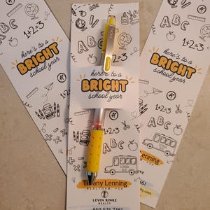 May include: Three white cards with black and yellow illustrations of school supplies and the text "here's to a BRIGHT school year". A yellow pen with a gold top is on the middle card. The bottom of the middle card has the text "Tivany Lenning REALTOR® PSA LEVIN RINKE REALTY 850.525.7461 tiffanyl@levinrinkerealty.com #propertybytiffany"