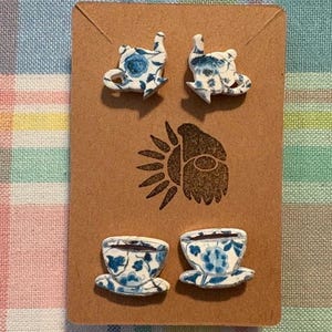 May include: A set of four blue and white ceramic earrings featuring teacups and teapots. The earrings are displayed on a brown card with a sun logo.