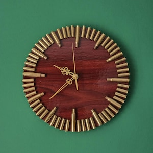 May include: A circular wall clock featuring a dark reddish-brown wooden face. The perimeter is decorated with a series of upright brass bullet casings arranged as hour markers. The clock has three thin, ornate gold-colored metal hands.