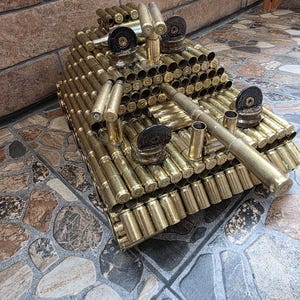 May include: A decorative model tank constructed entirely from metallic brass bullet casings. The structure features a central turret with a long barrel, layered cylindrical components forming the body, and circular dark metal accents on top.
