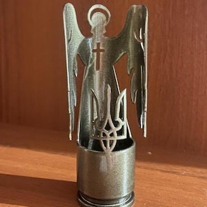 May include: A bronze-colored angel figurine with outstretched wings, a halo, and a cross. The angel's body features a Ukrainian trident symbol. The figurine is mounted on a cylindrical base, likely a repurposed bullet casing.