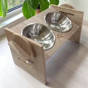 May include: A light brown wooden elevated pet food and water bowl stand with two stainless steel bowls. The stand has a paw print design.