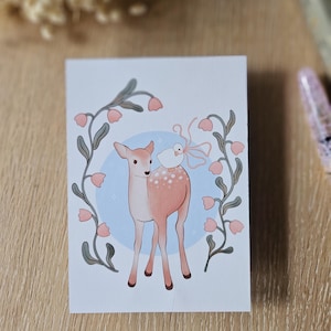 Peut inclure: Une impression d'art rectangulaire représentant un faon à taches blanches devant un cercle bleu clair. Un petit oiseau blanc avec un ruban rose est posé sur le dos du faon. La scène est encadrée par deux tiges verticales à fleurs roses.