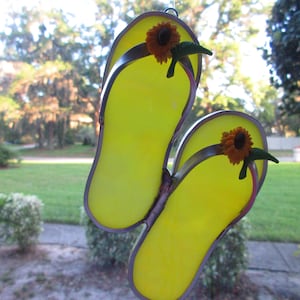 May include: A pair of yellow glass flip-flops with a sunflower on each strap. The flip-flops are hanging from a window.