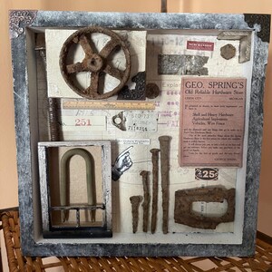 May include: A framed shadow box art piece with a rustic theme. The box contains various vintage hardware items, including a rusty gear, nails, a metal U-shaped piece, and a piece of paper with the text "GEO. SPRING'S Old Reliable Hardware Store Union City MICHIGAN".