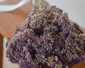 Puede incluir: Un ramo de flores moradas secas atado con una cinta de raso blanco. Las flores tienen un aspecto denso y texturizado. El ramo descansa sobre una superficie de madera, lo que sugiere un tema rústico o natural.