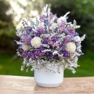 Peut inclure: Un bouquet vibrant de fleurs séchées dans un vase en céramique blanc. L'arrangement présente des fleurs violettes, blanches et lavande, créant un affichage texturé et coloré. Le vase est posé sur une surface en bois avec un arrière-plan vert flou.