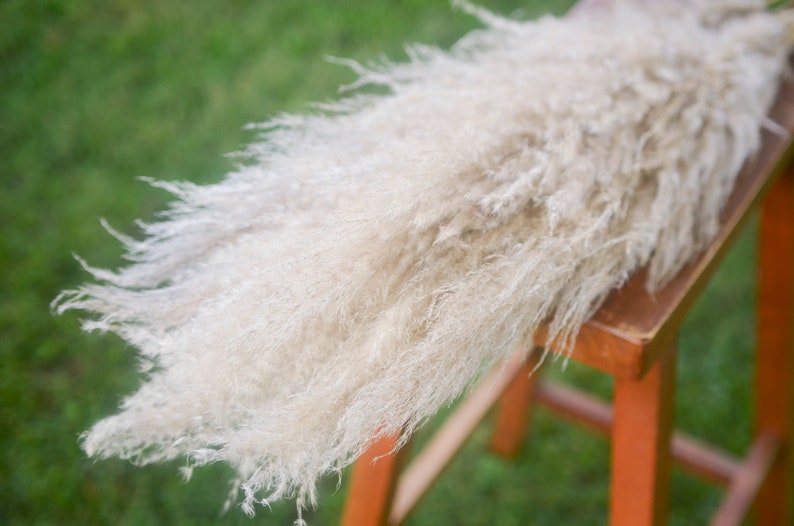 May include: A large bunch of dried pampas grass with fluffy, feathery plumes in a light beige color. The grass is arranged on a wooden stool with a bright orange finish.