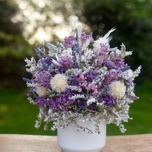 May include: A floral arrangement of dried purple lavender, white statice, and cream-coloured globe amaranth flowers displayed in a simple, smooth white ceramic pot. The bouquet features a dense mix of textures and shades of violet, lavender, and white.