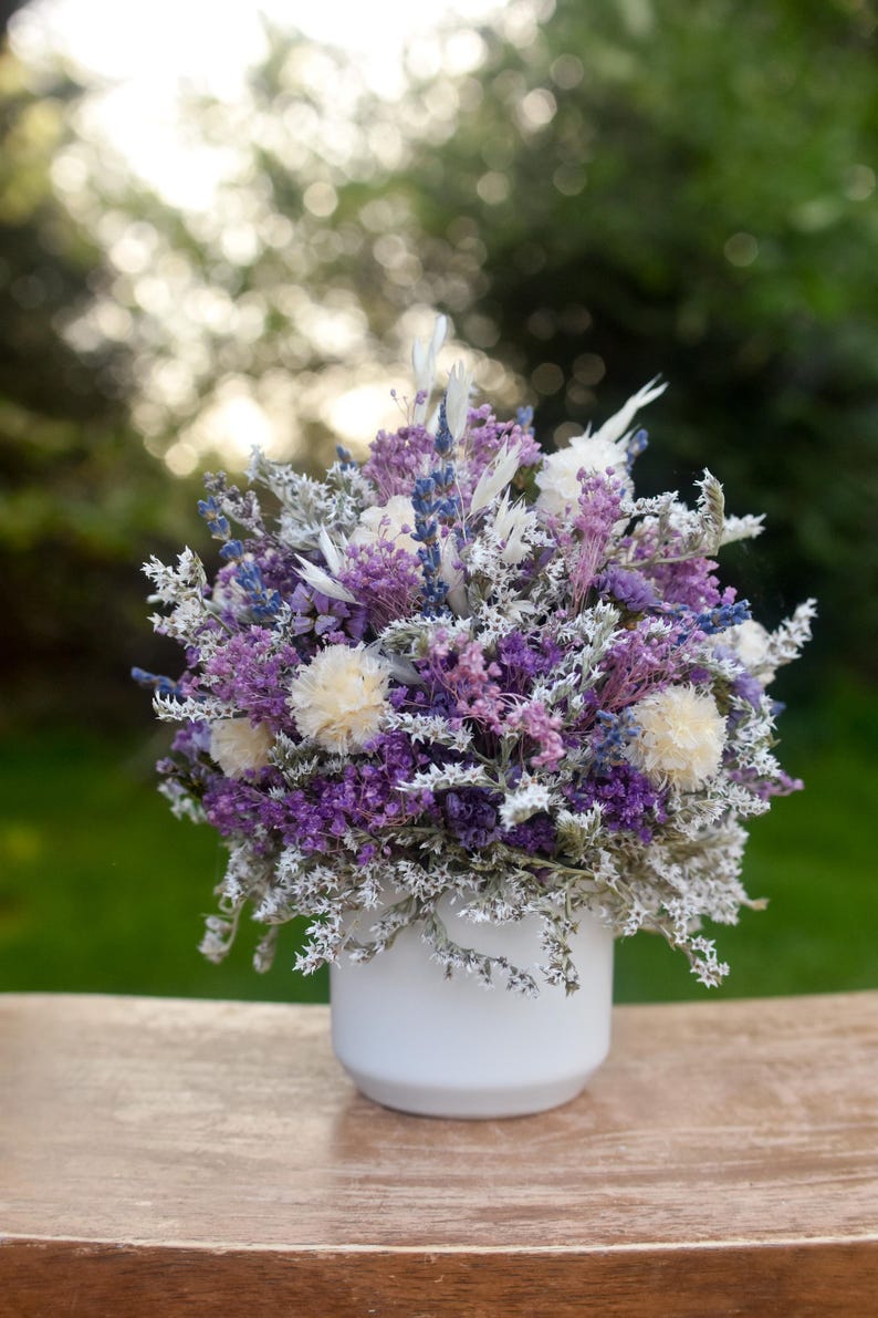 May include: A floral arrangement of dried purple lavender, white statice, and cream-coloured globe amaranth flowers displayed in a simple, smooth white ceramic pot. The bouquet features a dense mix of textures and shades of violet, lavender, and white.