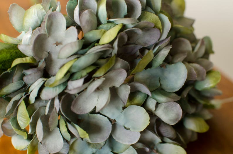 May include: Close-up of a bouquet of artificial hydrangea flowers. The petals are a mix of muted blue-green and gray, with hints of yellow-green. The arrangement is dense and full, suggesting a floral centerpiece or decorative element.