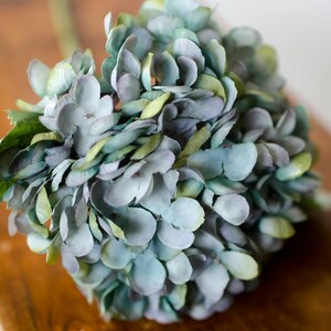 May include: A close-up of a faux hydrangea flower arrangement. The flower head is a mix of blue, green, and gray petals. The arrangement sits on a wooden surface. The stem and leaves are green.