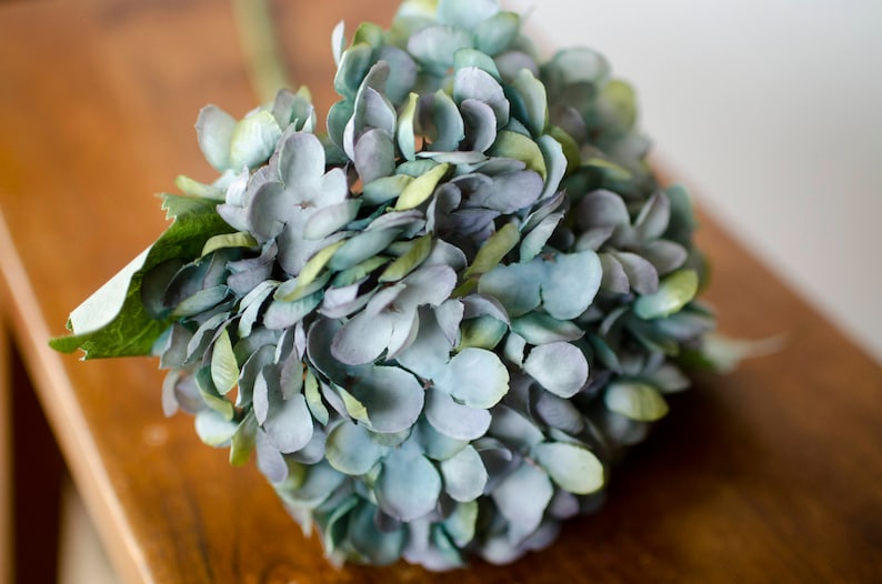 May include: A close-up of a faux hydrangea flower arrangement. The flower head is a mix of blue, green, and gray petals. The arrangement sits on a wooden surface. The stem and leaves are green.