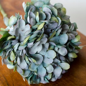 May include: A close-up of a faux hydrangea flower arrangement. The flower petals are a mix of blue, green, and purple hues. The arrangement is on a wooden surface, with the stem and leaves visible. The overall aesthetic is natural and decorative.