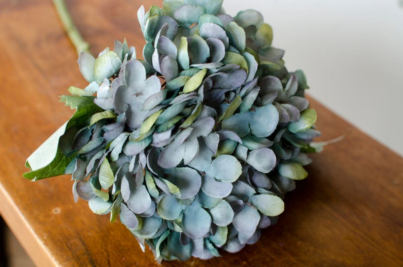 May include: A close-up of a faux hydrangea flower arrangement. The flower petals are a mix of blue, green, and purple hues. The arrangement is on a wooden surface, with the stem and leaves visible. The overall aesthetic is natural and decorative.