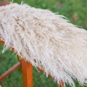 May include: A bunch of dried pampas grass with long, feathery plumes in a light beige color. The grass is arranged on a wooden stool.