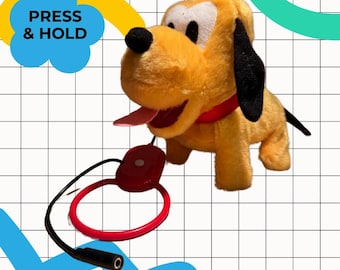 Peut inclure: Un jouet en peluche représentant un chien jaune avec des oreilles noires, un collier rouge et une langue rose. Il est équipé d'un bouton rouge et d'un cordon noir. Le texte "PRESS & HOLD" est visible.