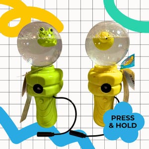 Pode incluir: Duas varinhas de bolhas portáteis com topos esféricos transparentes contendo líquido e pequenas figuras flutuantes. Uma varinha é verde-limão com um sapo, a outra é amarela com um pato. Ambas têm cordões pretos e uma etiqueta azul: PRESS & HOLD.